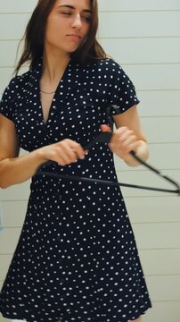 Caucasian woman dancing in polka dot dress, swinging hanger and spinning near mirror in lively fitting room, joyful movement, candid smile, stool and shopping bag in background, upbeat playful energy