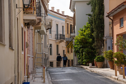 A ground-level view of a quiet, sun-drenched street in Plaka, Athens where two pedestrians walk past elegant neoclassical buildings.