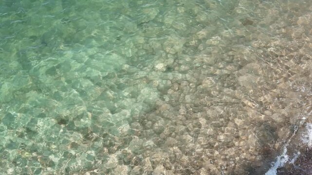 crystal clear turquoise sea water with small waves gently lapping onto a sandy pebble beach. Natural summer sunlight reflecting on the water surface. Refreshing vacation background.	
