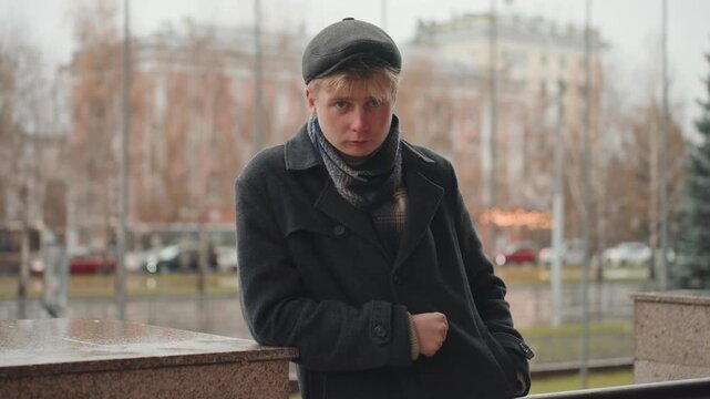 Wistful white creator leaning on stone railing, intimate closeup of young white male storyteller creator with scarf and cap, raindrops on fabric, park evergreens in background, muted palette, raw
