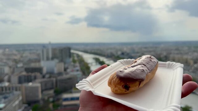 POV Point of view of hand holding a delicious chocolate eclair on a disposable plate capturing the beautiful cityscape of Paris, France, with the winding Seine River in the background