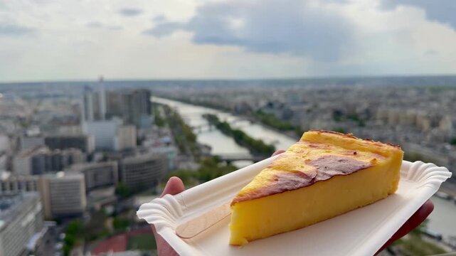 POV Point of view of hand holding a slice of French cheesecake on a disposable plate, enjoying a sweet treat with the panoramic view of the Paris cityscape and Seine River in the background