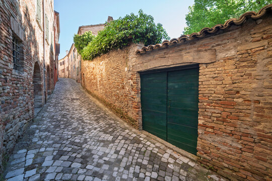 Narrow cobblestone street lined with old brick walls and a green wooden gate in Mondaino, Emilia Romagna, Italy, illuminated by soft morning light and framed by lush greenery