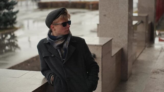 Stylish white creator leaning on railing overlooking rainy city plaza, hands in pockets with sunglasses and layered coat, reflective puddles and muted tones, cinematic street fashion scene perfect