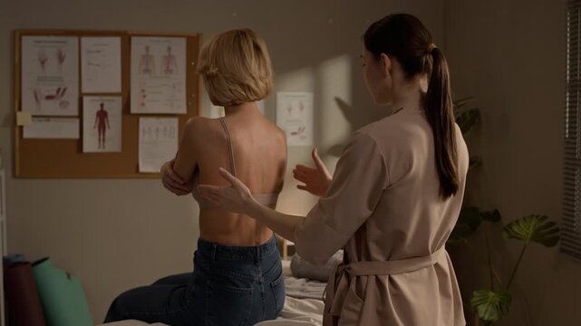 Back view medium shot of female therapist assessing client back muscles and posture before massage in cozy treatment room