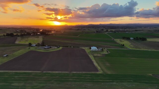 Aerial video panning over green and cultivated fields in Morpeth in NSW, Australia beneath colourful sunset sky. Scenic rural landscape showing farmland patterns and warm evening light 