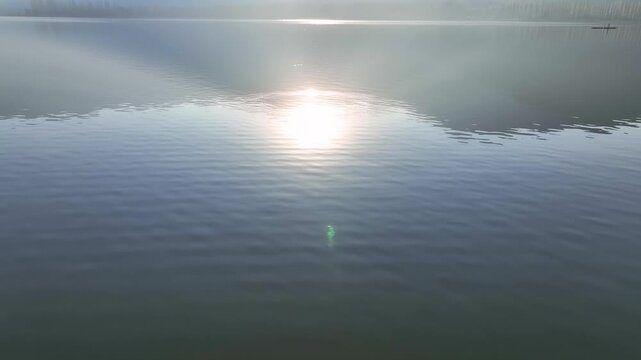Cinematic backward drone flight and tilt-up reveal starting from the calm waters of Dal Lake, transitioning up to a glorious sunrise and the dark silhouette of a mountain in Srinagar.