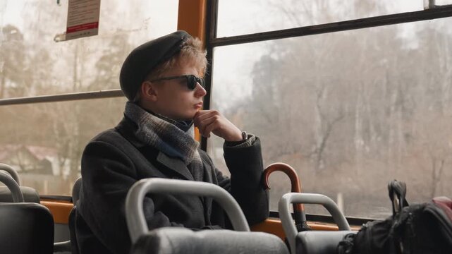 Young white creator on bus, gazing through window at autumn forest wearing scarf, cap, sunglasses hand near face, umbrella beside seat, backpack at feet cinematic moody portrait for storytelling