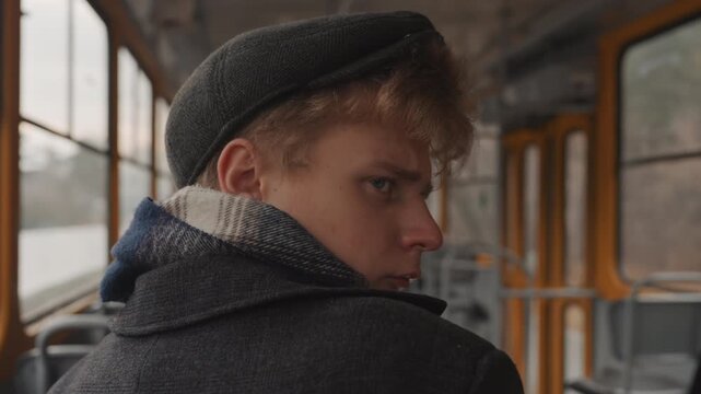 White creator on tram looking back, knitted cap and scarf, vintage interior, soft overcast light young white writer glances over shoulder with pensive expression, empty seats and window reflections