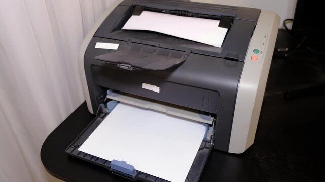 Close view of a laser printer feeding a white sheet, paper tray open on a dark desk in office light. Home printing supports paperwork and taxes today, but costs and waste need control.