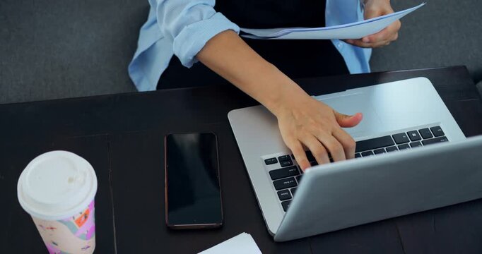 Hands typing on laptop, reviewing report and data sheets professional fingers move across keyboard while phone and coffee sit ready, flipping through paper pages to crosscheck figures, edit summary,