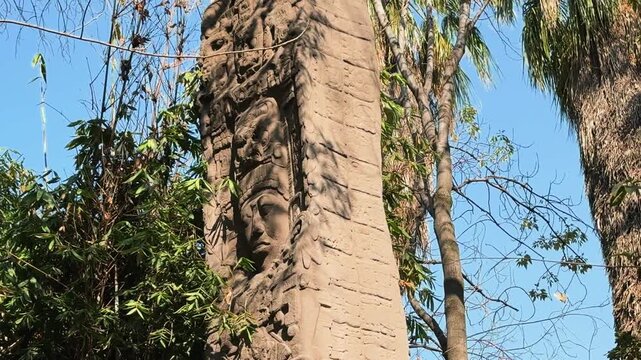 Towering stone column carved with ancient face relief at Museo Nacional de Antropolog&iacute;a in Mexico City. Pre-Columbian sculpture surrounded by tropical trees under blue sky.