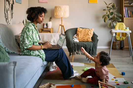 African American young mother working on laptop while toddler interrupting from floor at home, showing remote work and parenting balance