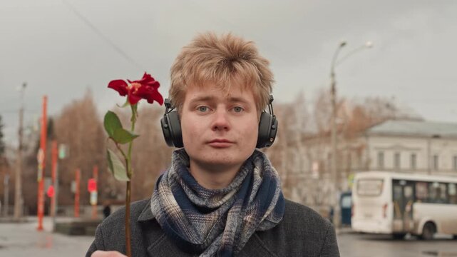 White creator with headphones holding rose, street poet offering flower then flipping finger, autumn city background, scarf and coat, playful smirk, candid urban portrait with cloudy sky and bus stop.