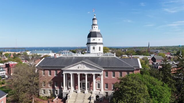Aerial drone view of Maryland State House in Annapolis, Maryland, USA during spring with blue sky, historic brick capitol building, white columns, dome architecture and Chesapeake Bay backdrop. Rising