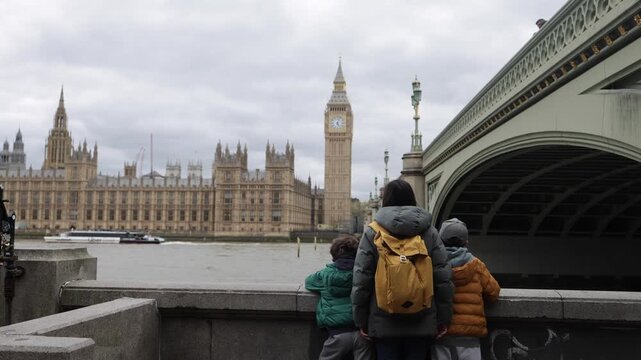 Mother and children watching big ben and the palace of westminster in london from across the river thames on an overcast day with a boat passing under westminster bridge