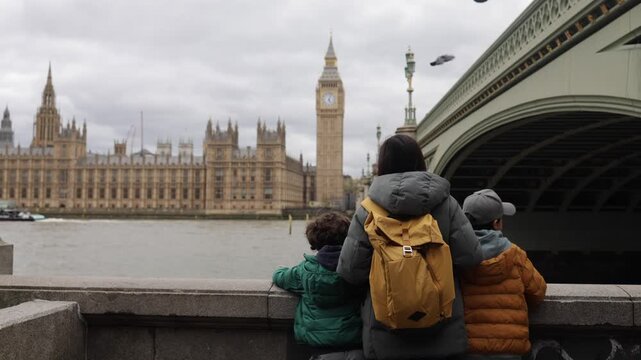 Mother and her two young sons admiring the majestic palace of westminster and the iconic big ben clock tower from across the river thames on a cloudy london day