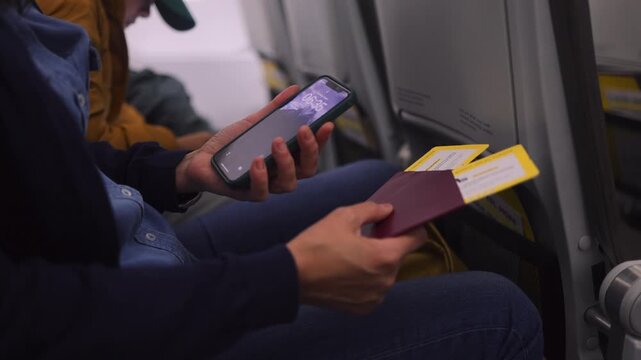 Woman passenger sitting in an airplane holding a passport, boarding passes, and a smartphone, getting ready for an international flight and checking the time