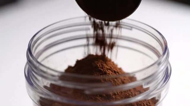 Powdered cocoa pouring into clear glass jar and plastic container, forming rich brown mound with fine particles and soft texture, showcasing smooth texture and warm atmosphere in close up view