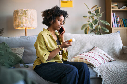 African American young adult woman holding prescription bottle while speaking on smartphone, managing treatment at home