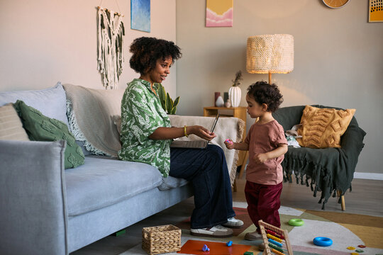 African American young mother working on laptop while caring for toddler at home, balancing parenting and remote job