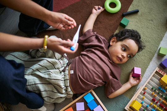 Overhead shot of toddler lying on play mat while parent checking thermometer, suggesting childhood fever and home care