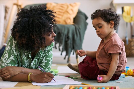 Black mother guiding toddler while drawing on paper during floor play at home, supporting early learning and bonding