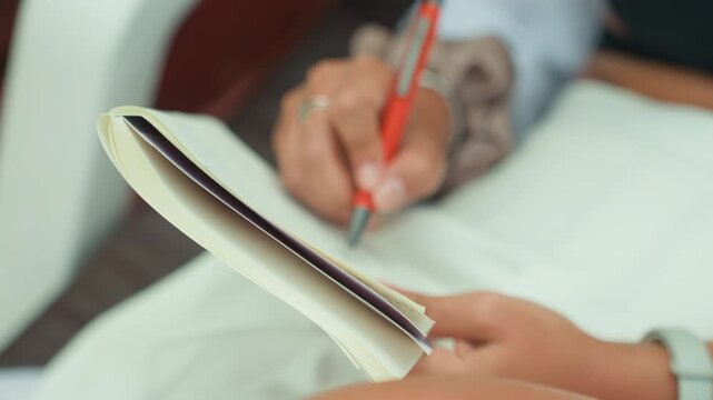 Close up hands writing island plan on lap, notebook pages slightly curled, red pen moving across lined paper, bracelet and watch visible, calm resort cabin mood, focused travel planning