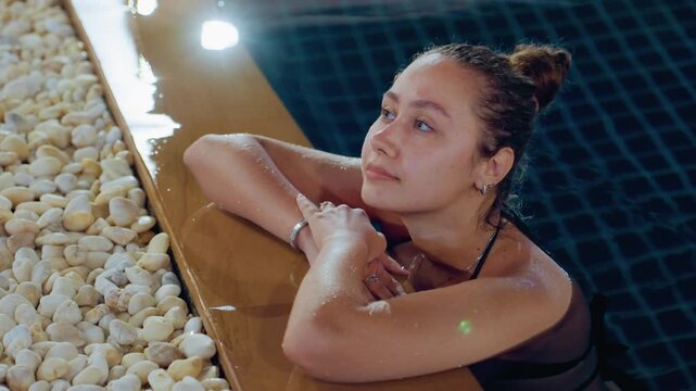 Night poolside woman leaning on wooden edge with pebble border at island resort, blue tiled water and lantern glow, wet shoulders and bun hairstyle, bracelets catching light, reflective gaze and quiet