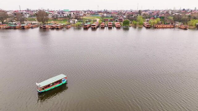 Cinematic aerial orbit shot of a traditional green-roofed shikara boat navigating the calm waters of Dal Lake, with a backdrop of floating houseboats and Himalayan mountains in Srinagar