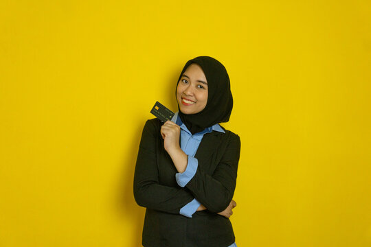 A woman wearing a hijab and business attire holds a credit card in front of a bright yellow background with a confident expression