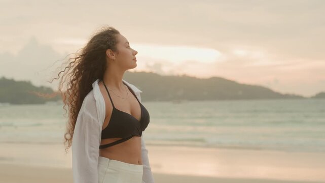 Woman on island beach at sunset, curlyhaired woman in black bikini and white shirt strolls along shoreline, pauses to gaze toward distant islets and kite, hands clasping shirt, soft golden light,