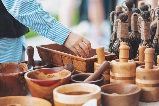 A person reaches into a wooden bowl at a craft fair. Wooden bowls, utensils, and other items are displayed on a table. People walk in the background under the sunlight
