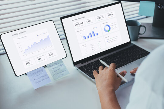 Remote work analysis scene, professional reviewing performance metrics on laptop and tablet displaying business dashboard, analytics charts, data insights in modern workspace decision making workflow