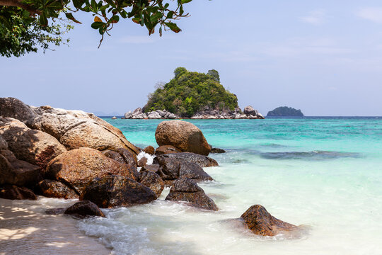 Ko Usen Islet view from Ko Lipe. Turquoise waters meet rocky shore under clear sky. Tropical trees frame the beach scene.