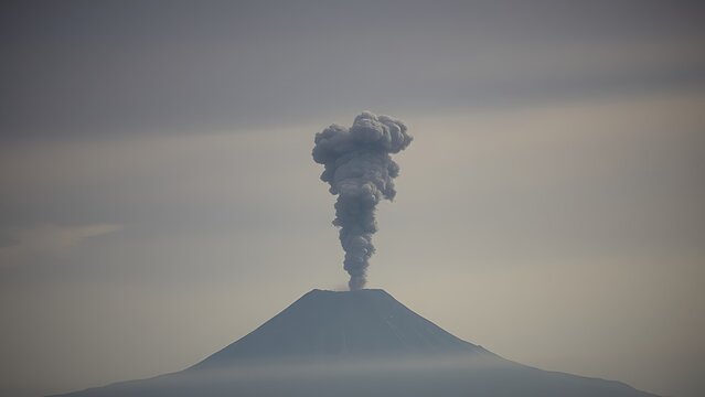 A distant volcano erupting, plume of dark smoke and ash in a grey sky.