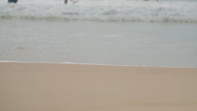 Calm island beach shore, gentle waves lapping pale sand, distant swimmers blurred on horizon, overcast light casting muted tones, soft foam edges, slow motion ebb and flow, minimal coastal mood