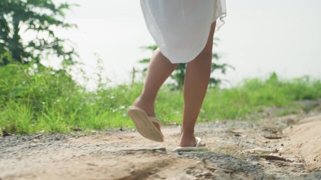 Lowangle legs walking along island shore, grassy verge and distant water, sandals scuff pebbles and dirt, purposeful exploratory stride, sunlit horizon and windswept greenery evoke adventurous seaside
