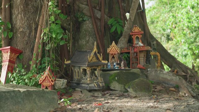 Miniature shrine at banyan tree, weathered spirit houses and offerings beneath massive roots, mosscovered stones, incense sticks, devotional figurines, ornate gilded roofs, quiet forest atmosphere,
