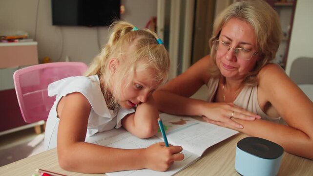 Girl doing school assignment at home desk with pencil, mother help learning process. Primary education concept