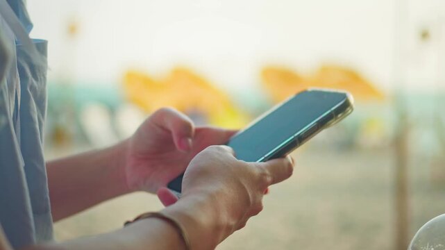 Close up hand of woman typing on smartphone at sandy beach. Woman hand text mobile phone on a sunny day at windy ocean side enjoy spend time on holiday vacation trip at the beach.