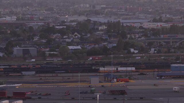 Aerial drone shot capturing the sprawling industrial landscape and freight train yard of Oakland, California, with distant city traffic moving during the hazy light of an early sunrise