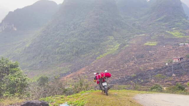 Motorcycle loaded with red touring gear parked on grassy roadside overlooking steep terraced mountainside with misty karst landscape in Dong Van Karst Plateau Geopark Ha Giang Northern Vietnam