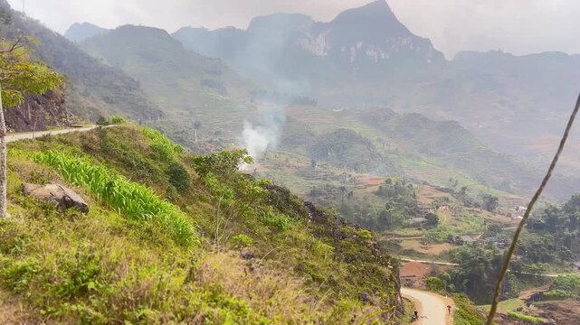 Winding cliffside road overlooking misty valley with terraced fields and rising smoke in Ma Pi Leng Pass Ha Giang Loop Northern Vietnam showing rugged terrain and rural life