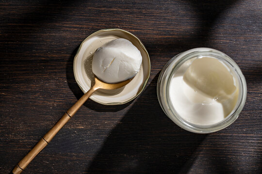 Top view of fresh thick organic yogurt or creamy soft cheese or sour cream in a glass jar with a golden spoon on dark wooden background with dramatic shadows