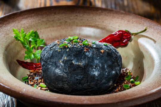 Whole potato roasted in charcoal ash served in a rustic ceramic bowl with green onions, herbs and chili peppers, closeup. Modern gourmet cuisine and creative food photography