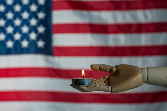 Symbolic image of a wooden mannequin hand holding a burning candle against blurred American flag. Concept for national mourning memorial day prayer and patriotism