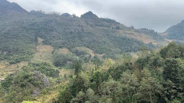 Mountain road along hillside overlooking green karst valley with scattered homes and forested slopes in Dong Van Karst Plateau Geopark Ha Giang Northern Vietnam under cloudy sky