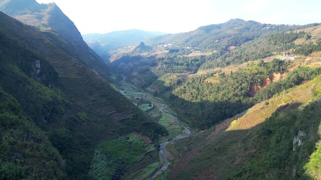Aerial drone shot of winding road snaking through rocky terraced valley with river below under hazy sky in Dong Van Karst Plateau Geopark Ha Giang Northern Vietnam