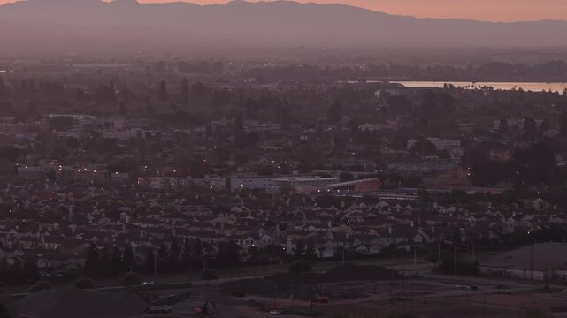 Tracking aerial view from a drone flying over Alameda during a hazy sunrise, with distant mountains and city lights visible through the morning mist